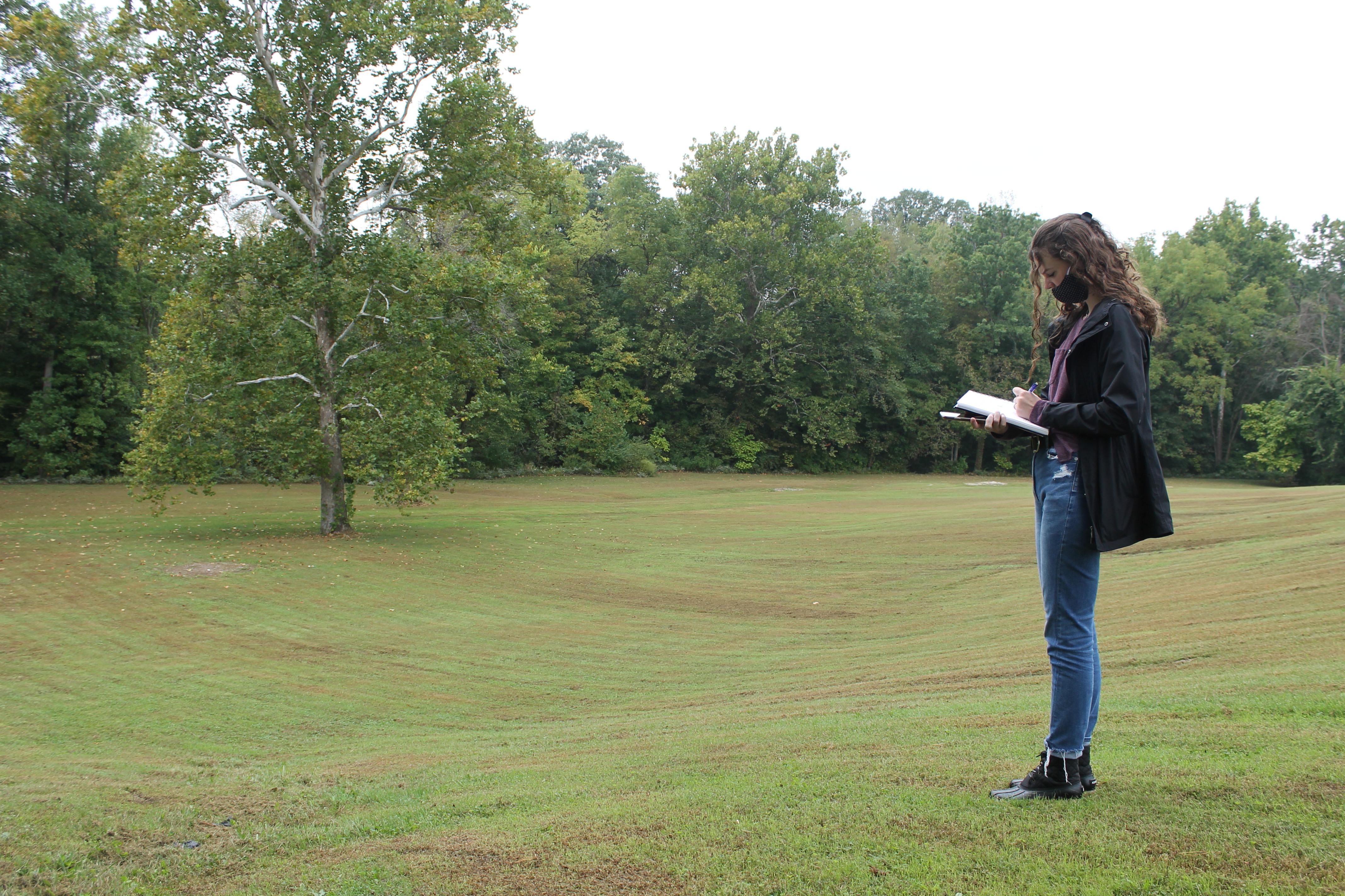 一个长着棕色头发，戴着面具的学生站在图像的左边，面向右边. 她身后是一片树木覆盖的空地.